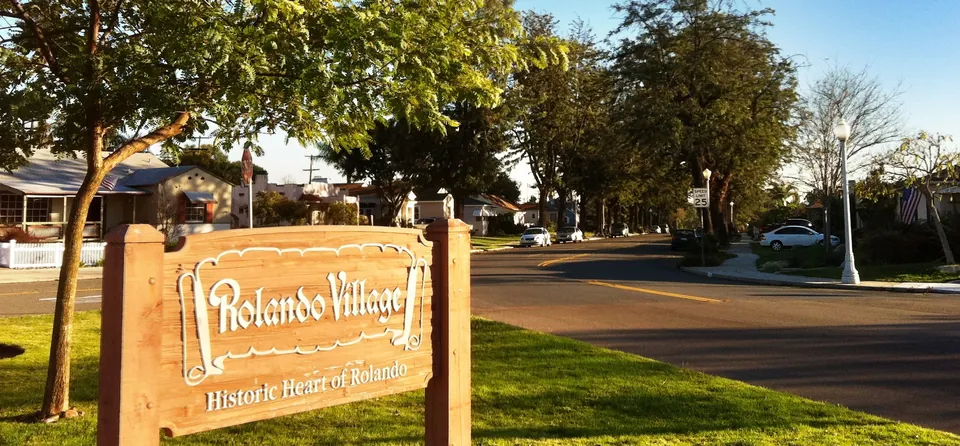 Carved “Rolando Village — Historic Heart of Rolando” sign beside a tree-lined Rolando Boulevard